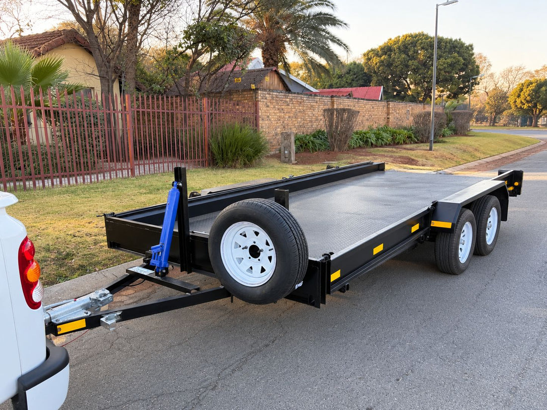 Black double-axle flat-deck trailer with a spare wheel, hitched to a white bakkie